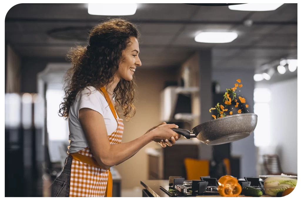 Mulher em cozinha salteando legumes. 